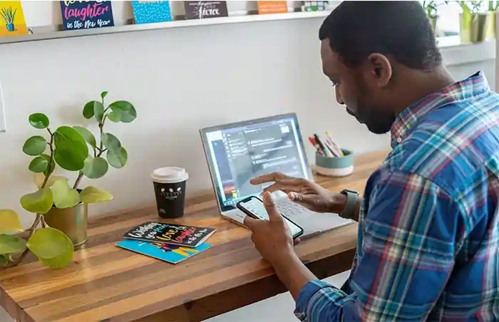 A person holding a phone and looking at a laptop screen, utilizing the Multi-User Chat Support feature in DatumOffice
